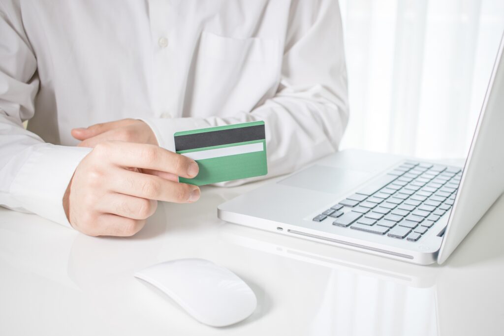 A person holding a green credit card with a laptop and a computer mouse on a white table